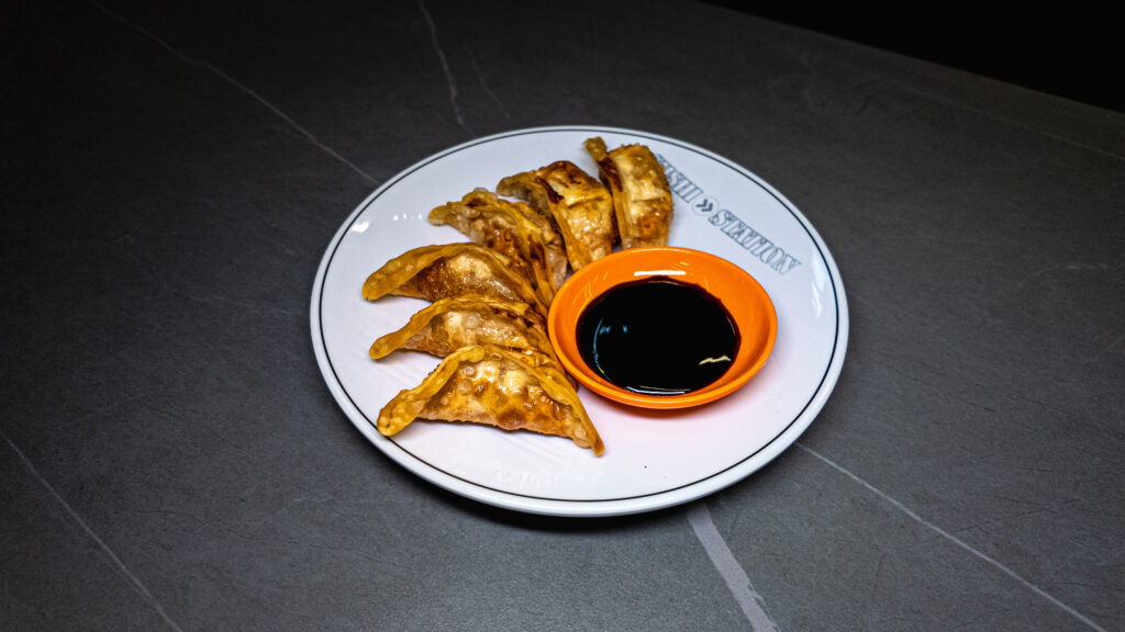 Crispy pan-fried gyoza dumplings served on a Sushi Station plate with soy sauce in an orange bowl, ready to enjoy at the W...