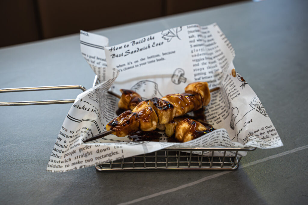 Grilled skewers with caramelized glaze served in a metal basket lined with sandwich-themed paper at Sushi Station in Westf...
