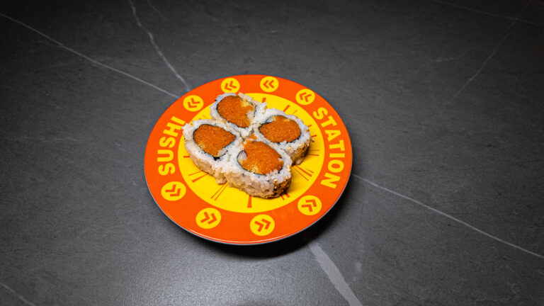 Four pieces of tobiko-topped sushi on a bright orange SUSHI STATION plate, set against a dark gray surface.