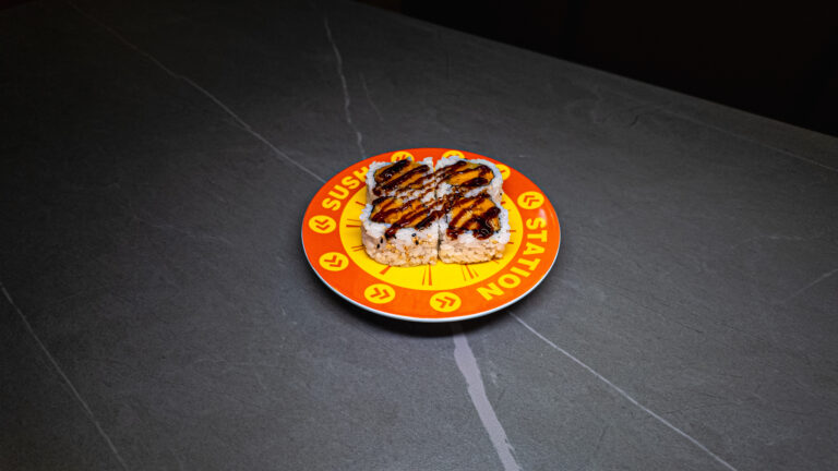 Four eel sushi rolls drizzled with sauce, served on a bright orange Sushi Station plate atop a dark stone table.