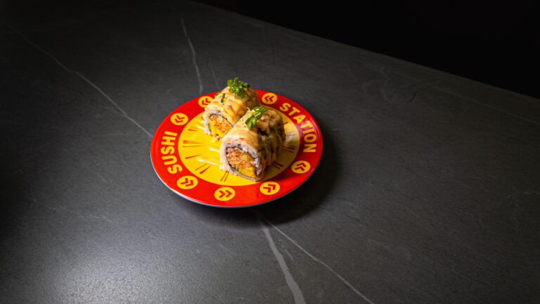 Sushi Station’s signature roll on a red “Sushi Station” plate, drizzled with sauce and garnished with green onions, set on...