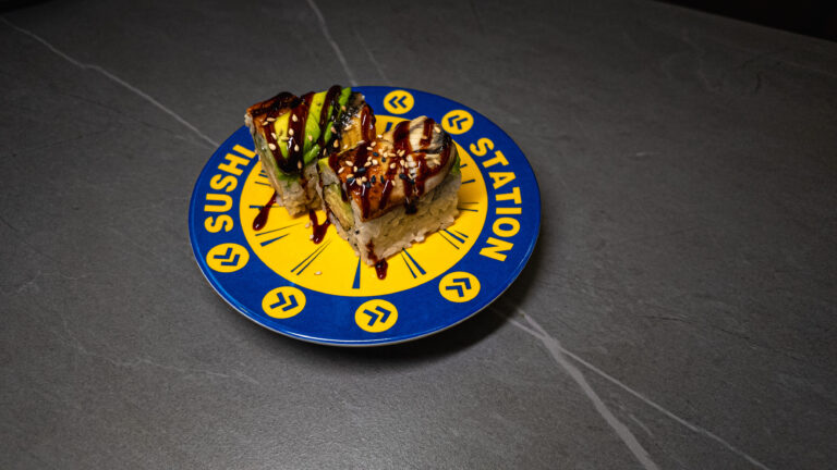 Sushi rolls with avocado and eel, drizzled with sauce and sesame seeds, served on a blue and yellow “Sushi Station” plate....