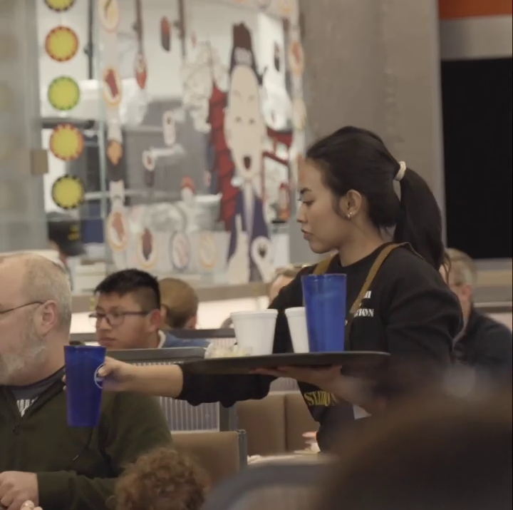 Waitress at Sushi Station in Westfield, IN, carries tray with blue and white cups through busy dining area with colorful m...